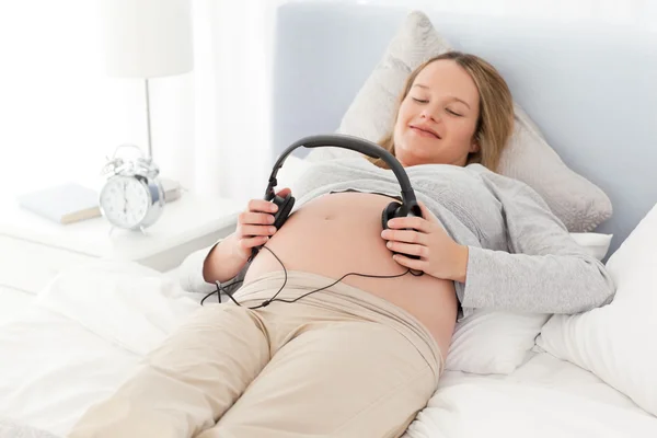Sorrindo futura mãe colocando fones de ouvido em sua barriga deitado em um ser — Fotografia de Stock