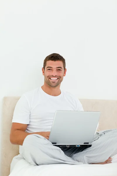 Homem alegre trabalhando em seu laptop sentado em sua cama — Fotografia de Stock