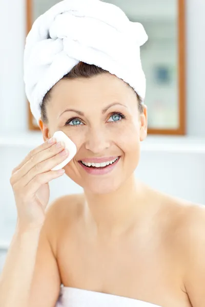 Mujer joven sonriente con una toalla poniendo crema en su cara en th Imagen de archivo