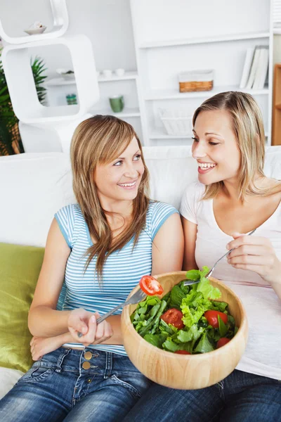 Two female friends eating salad together on a sofa Stock Picture