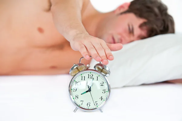 眠っている若い男が寝室で鳴っている目覚まし時計を停止します。 ストック画像