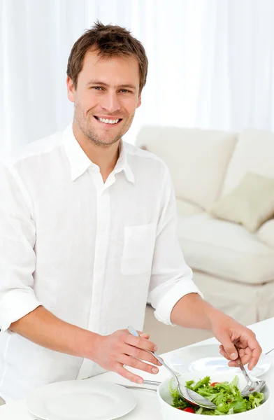 Hombre alegre sirviendo ensalada de pie en una mesa en la sala de estar — Foto de Stock