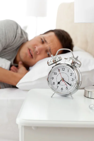 Homem infeliz olhando para seu despertador enquanto deitado em sua cama — Fotografia de Stock