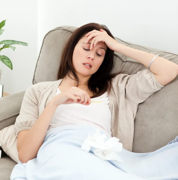 Mujer enferma mirando un termómetro mientras descansa en el sofá —  Fotos de Stock