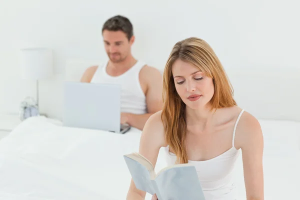 Мужчина сидит за ноутбуком, пока его жена читает книгу. — стоковое фото
