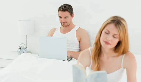 Мужчина сидит за ноутбуком, пока его жена читает книгу. — стоковое фото