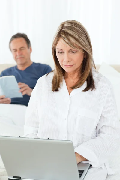 Woman looking at her laptop while her husband is reading — Stock Photo, Image