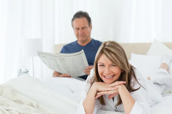 Usmívající se žena dívá na kameru, zatímco její manžel je readin — Stock fotografie