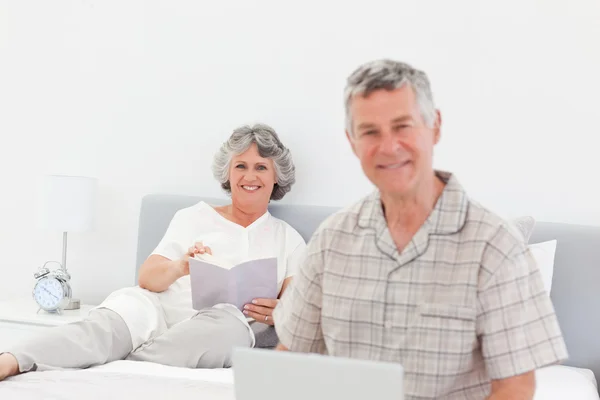 Man som arbetar på sin bärbara dator medan hans fru läser — Stockfoto