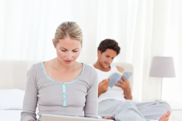 Woman working on her laptop while her husband is reading — Stock Photo, Image