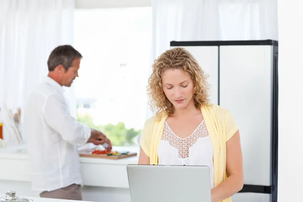 Mulher trabalhando em seu laptop enquanto seu marido está cozinhando — Fotografia de Stock