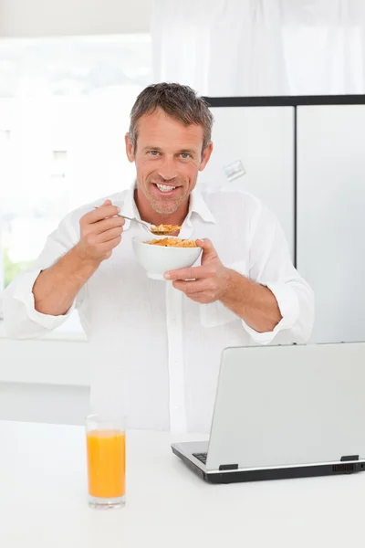 Homem comendo cereais enquanto ele está trabalhando em seu laptop — Fotografia de Stock