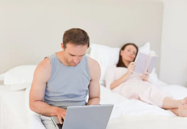 Manworking στο φορητό του ενώ η σύζυγός του διαβάζοντας ένα βιβλίο σχετικά με την — Φωτογραφία Αρχείου