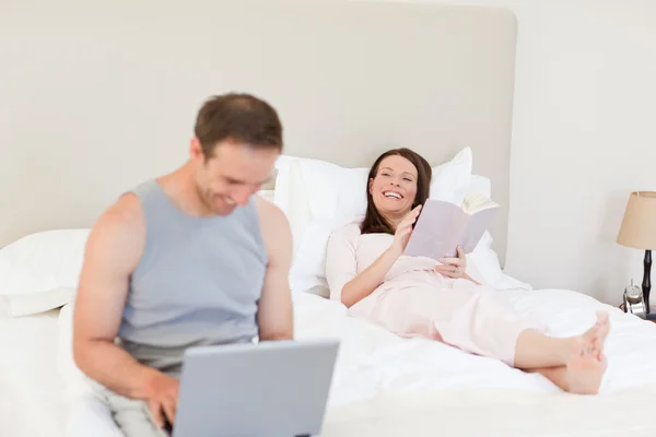 Работает над своим ноутбуком, пока его жена читает книгу о — стоковое фото