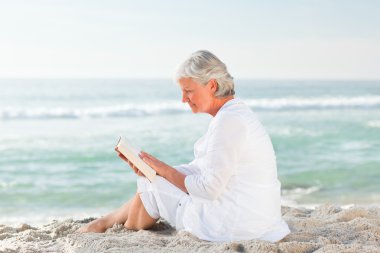 Elderly woman reading her book on the beach clipart