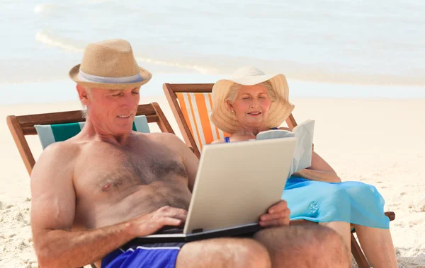 Hombre trabajando en su portátil mientras su esposa está leyendo en la playa — Foto de Stock