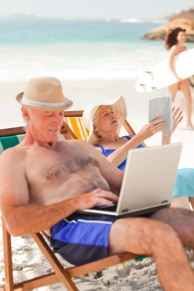 Hombre trabajando en su portátil mientras su esposa está leyendo en la playa — Foto de Stock