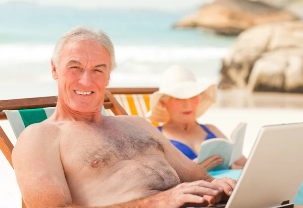 他的笔记本上工作，而他的妻子正在读取在海滩上的人 — 图库照片