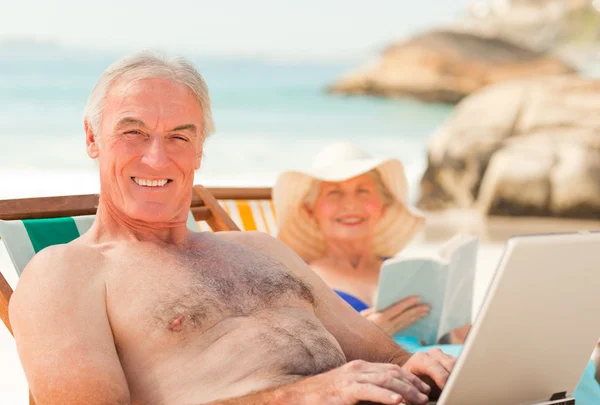 Man som arbetar på sin bärbara dator medan hans fru är att läsa på stranden — Stockfoto