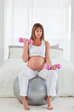 Beautiful pregnant female using a dumbbell while sitting on a gy clipart