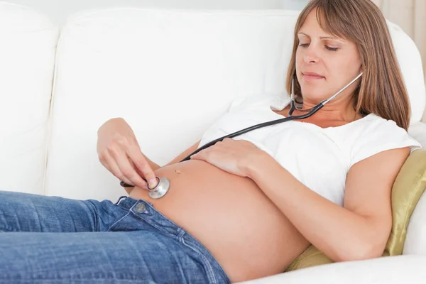 魅力孕妇躺在沙发上时使用听诊器 — 图库照片