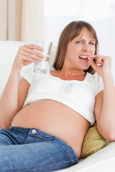 Mujer embarazada de buen aspecto tomando una píldora mientras está acostada en un sofá —  Fotos de Stock