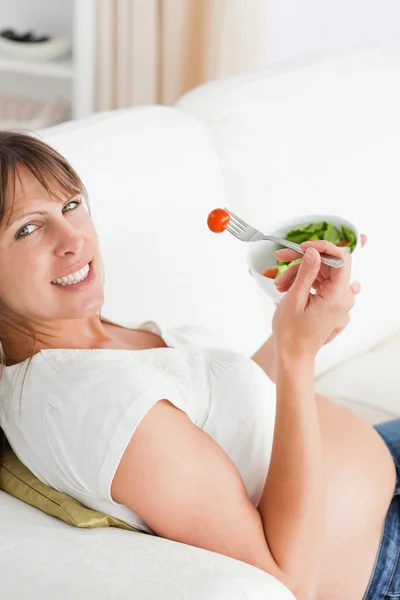 Ελκυστική έγκυος γυναίκα τρώει μια σαλάτα, ενώ βρίσκεται σε έναν καναπέ — Φωτογραφία Αρχείου