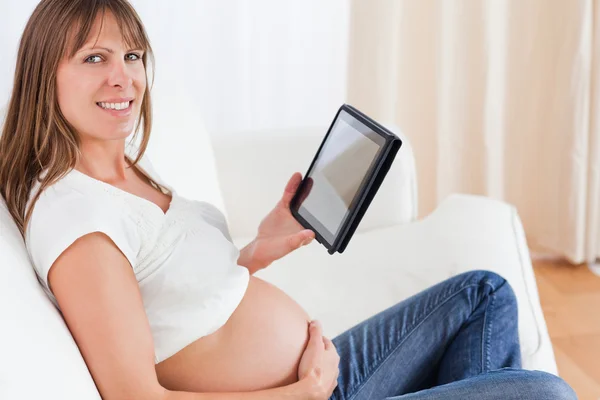 Encantadora mujer embarazada que se relaja con una tableta de ordenador mientras si — Foto de Stock