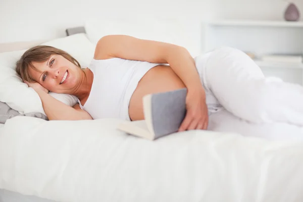 Linda mujer embarazada leyendo un libro mientras está acostada en su cama — Foto de Stock