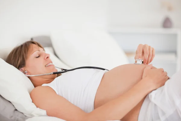 Милая беременная женщина, которая лежала на кровати, используя стетоскоп. — стоковое фото