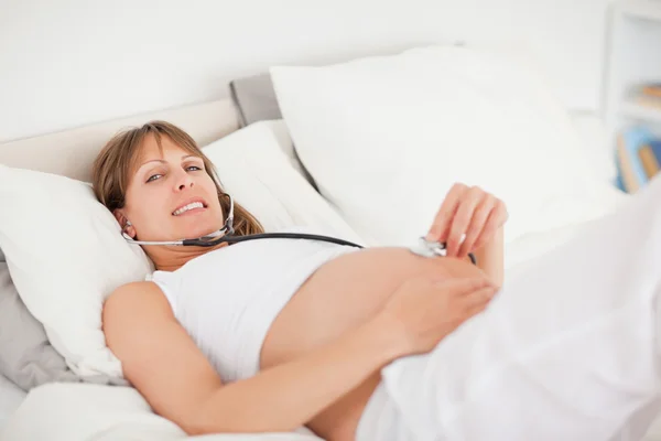 Очаровательная беременная женщина при помощи стетоскопа лежит на кровати — стоковое фото