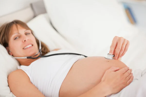 Mujer bastante embarazada usando un estetoscopio mientras está acostada en una cama — Foto de Stock