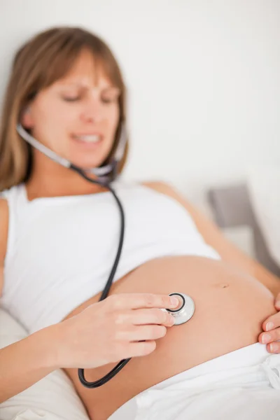 Aantrekkelijke zwangere vrouw met behulp van een stethoscoop terwijl liggend op een b — Stockfoto