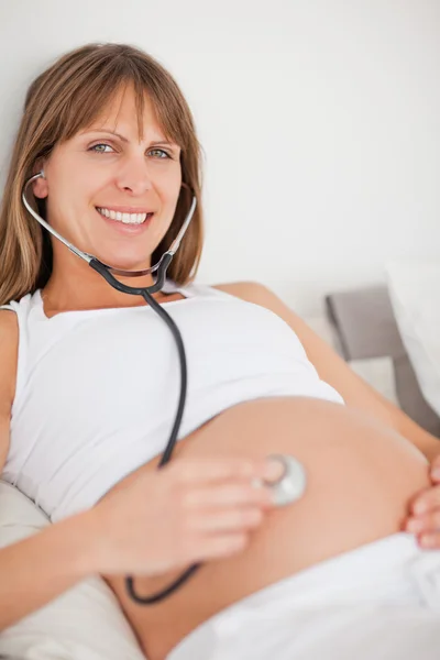 Bella donna incinta che utilizza uno stetoscopio mentre sdraiato su un letto — Foto Stock