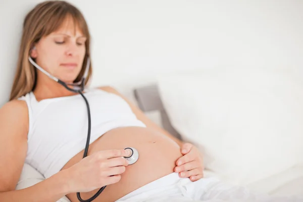 Belle femme enceinte en utilisant un stéthoscope tout en étant couché sur un be — Photo
