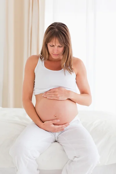 Прекрасная беременная женщина ласкает живот, сидя на кровати. — стоковое фото