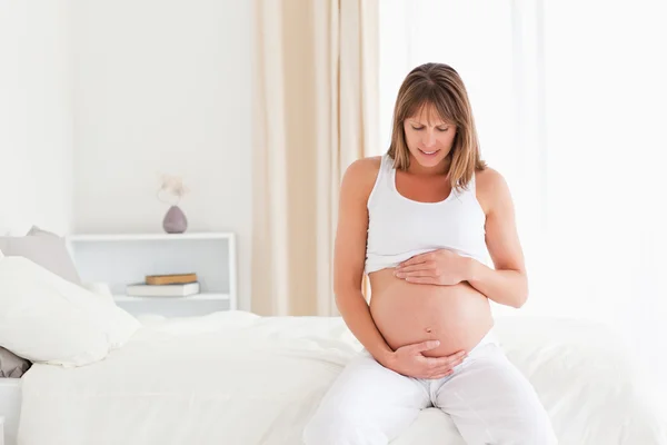 Atractiva mujer embarazada acariciando su vientre mientras está sentada —  Fotos de Stock