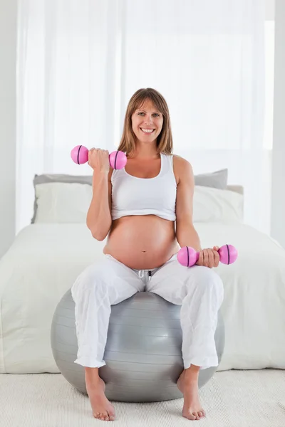 Atractiva hembra embarazada usando una mancuerna mientras está sentada en una g — Foto de Stock