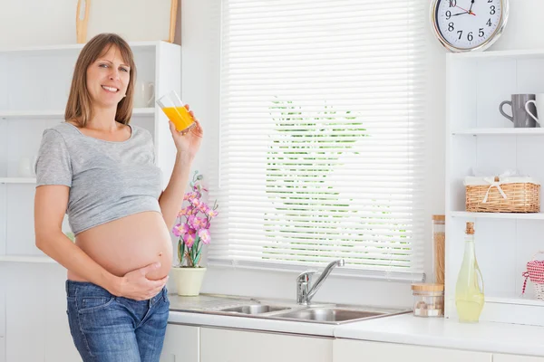 Donna incinta attraente che tiene un bicchiere di succo d'arancia mentre — Foto Stock