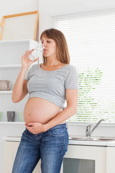 Mulher grávida de boa aparência segurando um copo de água enquanto está em pé — Fotografia de Stock