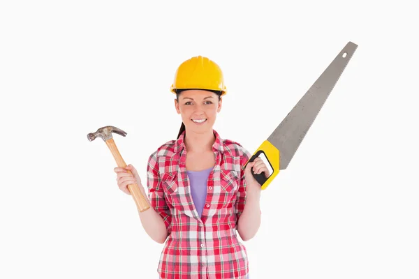 Mulher atraente segurando uma serra e um martelo enquanto está de pé — Fotografia de Stock