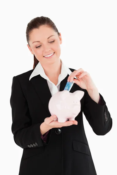 Mulher bonita em terno inserindo uma conta de dinheiro em um rosa porquinho b — Fotografia de Stock