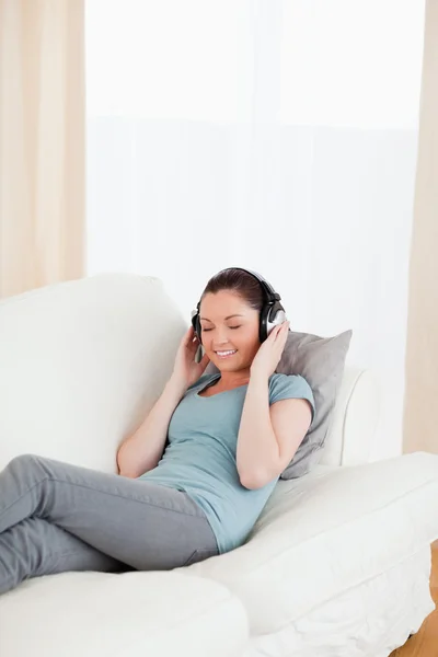 Ελκυστική γυναίκα χρησιμοποιώντας ακουστικά ενώ βρίσκεται σε έναν καναπέ — Φωτογραφία Αρχείου