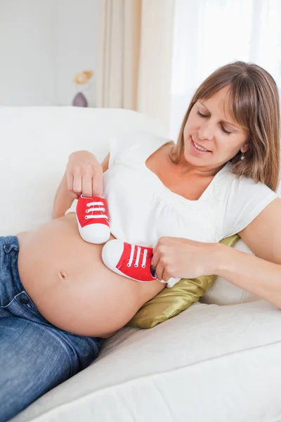 Charming pregnant woman playing with red baby shoes while lying — Stock Photo, Image