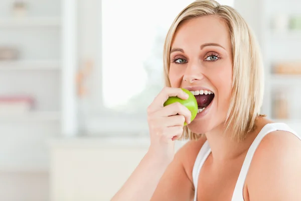 Young blonde woman sitting at table eating a green apple while l — Stock Photo, Image