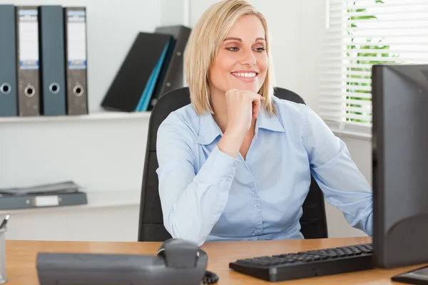 Lächelnde Frau mit dem Kinn in der Hand hinter einem Schreibtisch, der auf den Bildschirm blickt — Stockfoto