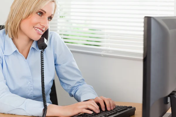 Smiling blonde businesswoman on the phone while typing looks at — Stock Photo, Image