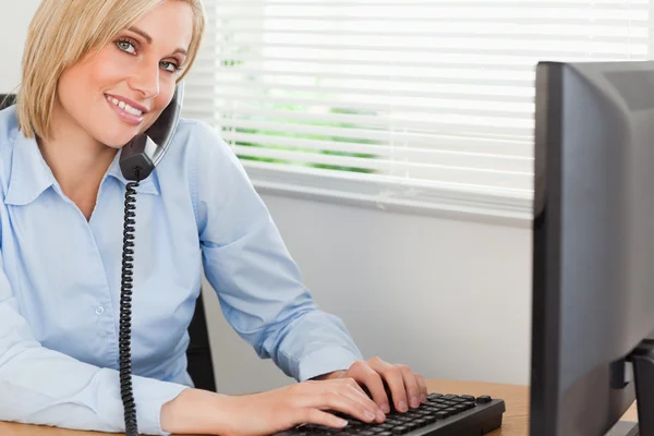 Smiling blonde businesswoman on the phone while typing looks int — Stock Photo, Image