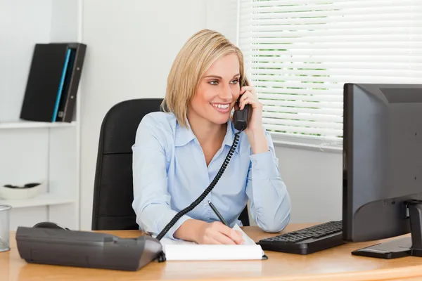 Cute businesswoman on phone writing something down looks at scre — Stock Photo, Image
