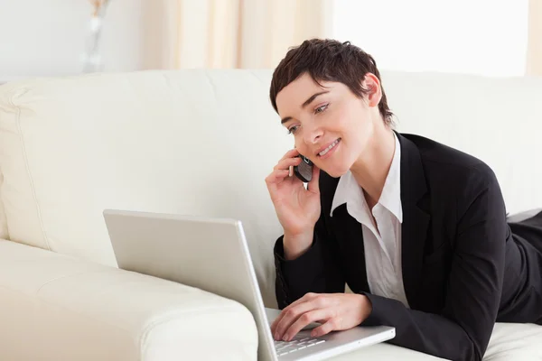 Mujer sonriente acostada en un sofá con un ordenador portátil y un teléfono — Foto de Stock